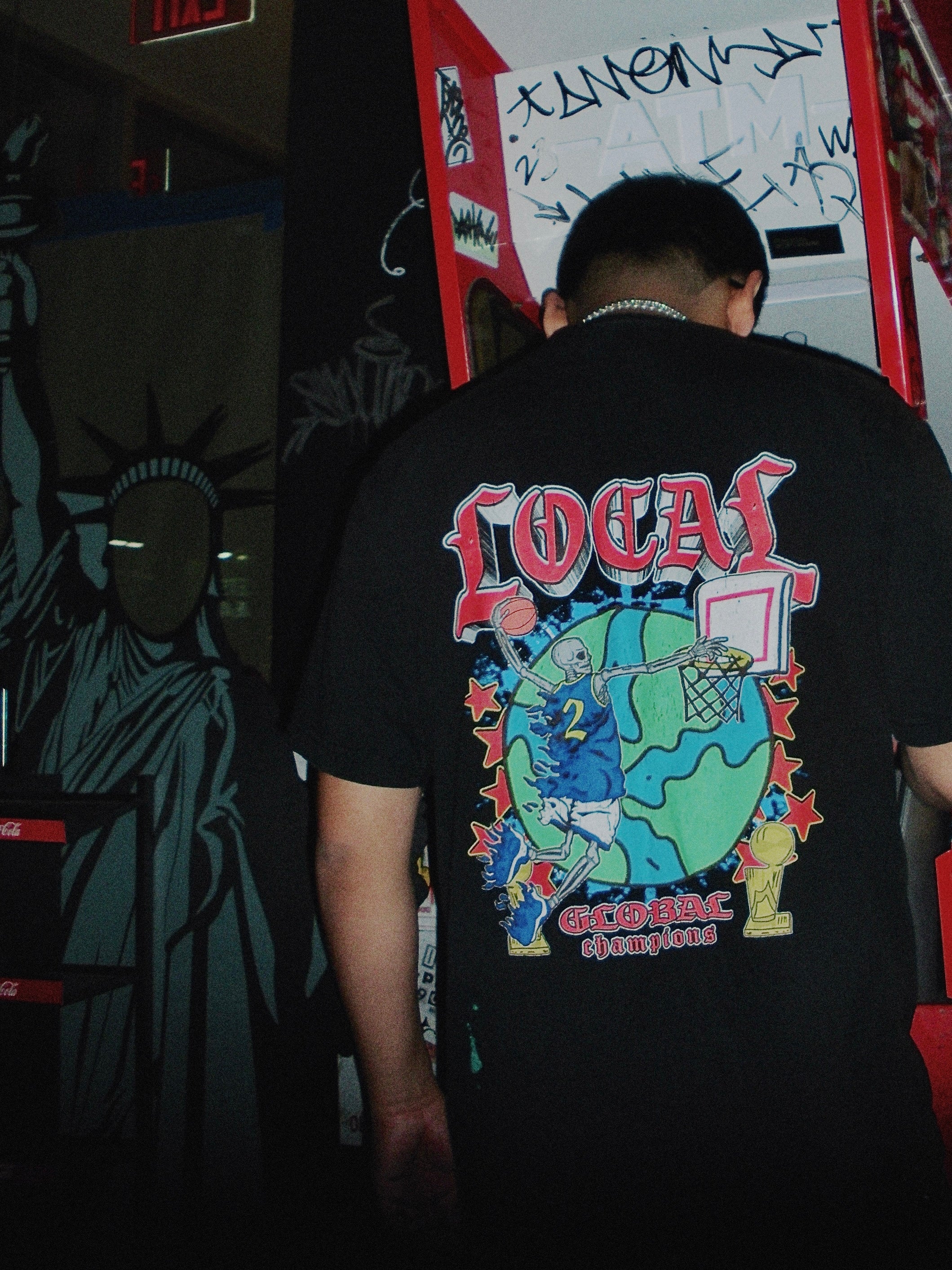 Person wearing a black t-shirt with colorful graphics and text, standing in a dimly lit room with graffiti on the wall.